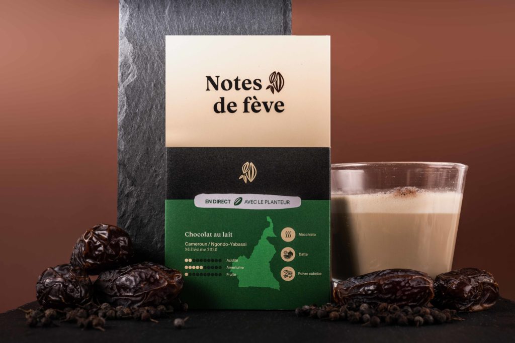 Une tablette de chocolat dans un emballage vert. Autour, un verre de Latte macchiatto, du poivre, et des dattes.